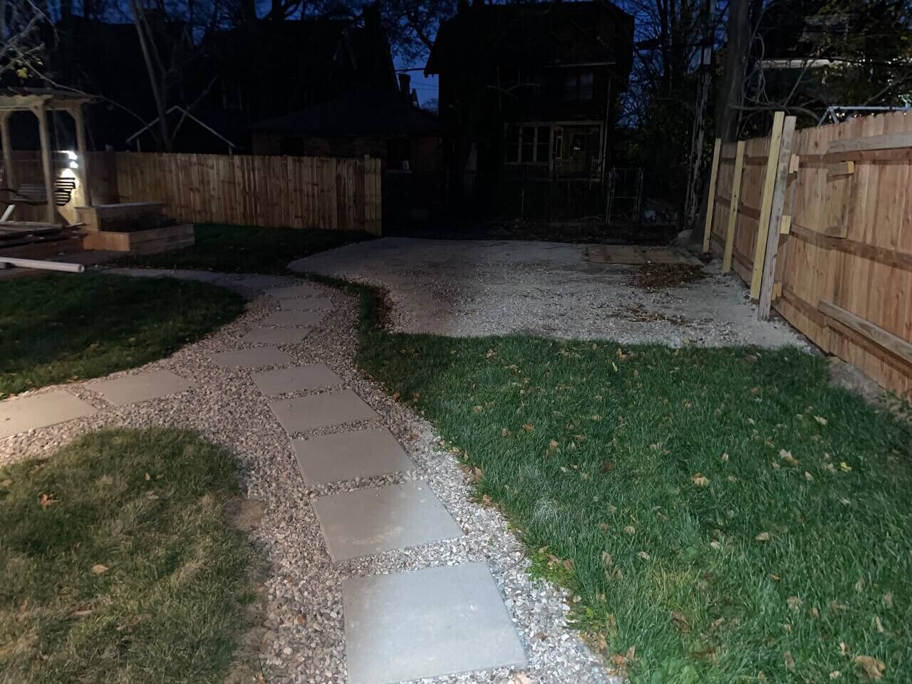 A paved pathway in a backyard with a wooden fence and a house in the background.