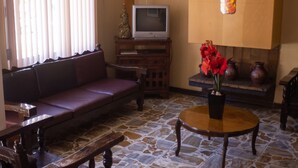 Una sala de estar con un banco de madera, una mesa de centro de madera, un televisor sobre un mueble de madera y un jarrón con flores rojas sobre una mesa de madera.