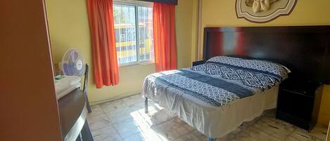 A bedroom with a bed, a desk, a chair, a window with curtains, a ceiling fan, and a decorative angel wall piece.