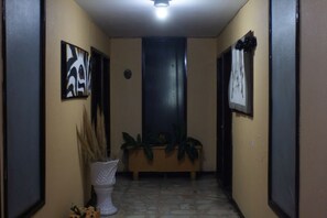 Un pasillo con una puerta oscura, un jarrón blanco con plantas secas y dos cuadros en las paredes.