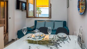 Un comedor con una mesa dispuesta para cuatro, un sofá con cojines estampados, un televisor en la pared y vista al mar por una ventana.
