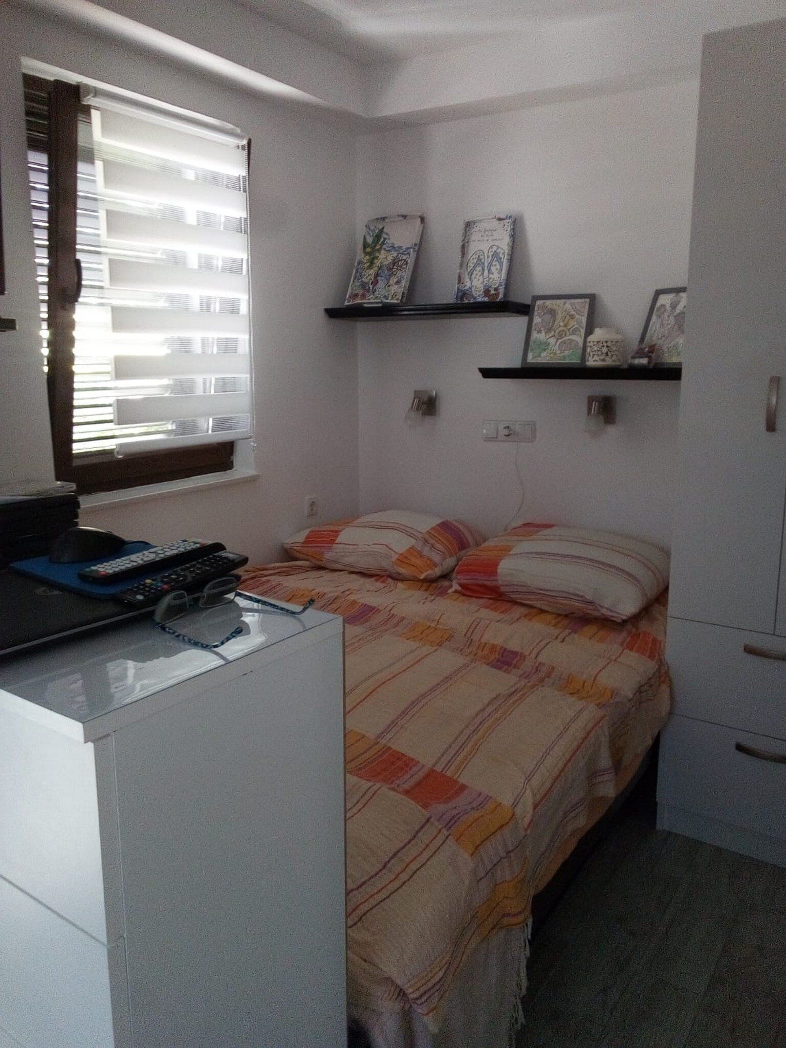 A small, neatly arranged bedroom with a bed, a desk, and a shelf with decorative items.