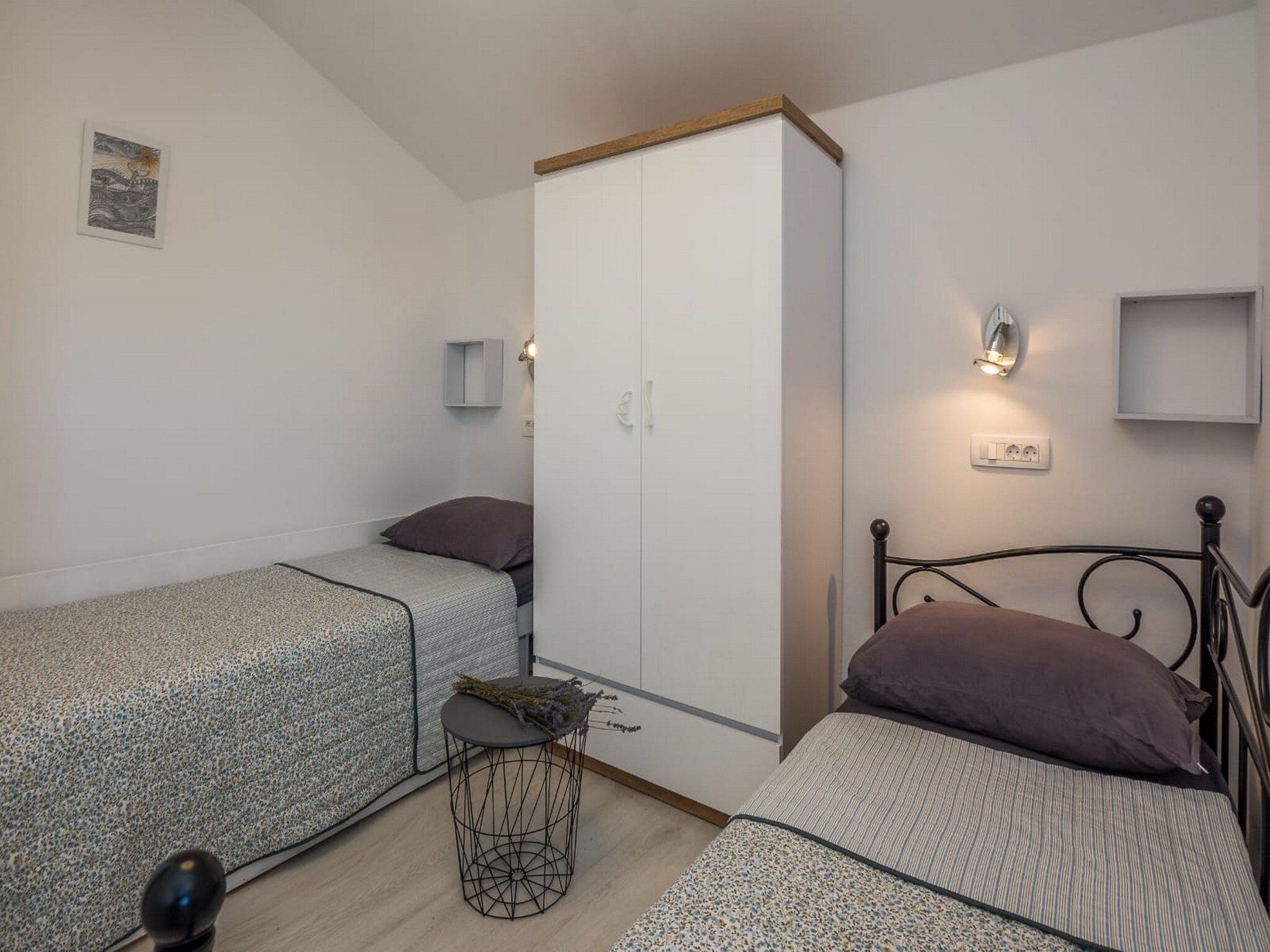 A small, well-lit room with two single beds, a wardrobe, a small round table, and a wall-mounted light fixture.