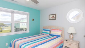 A bedroom with a bed, a window, a ceiling fan, a wall-mounted picture, a round mirror, and a bedside table with a lamp. - Aint Dat A Beach Entire - 3BR Home on the 2nd Row (North Topsail Beach)