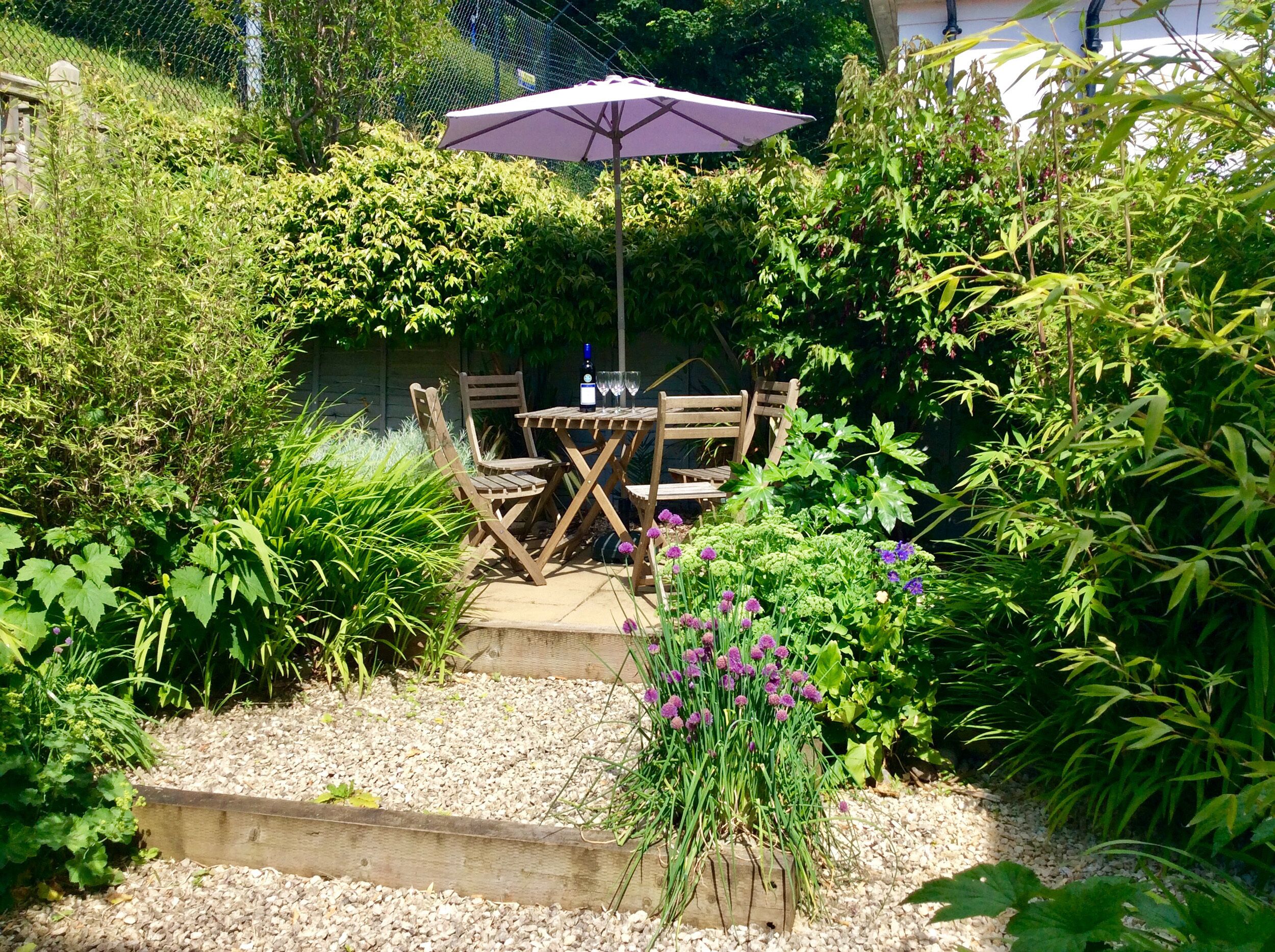 A garden patio with a table and chairs, surrounded by lush greenery and a gravel path.