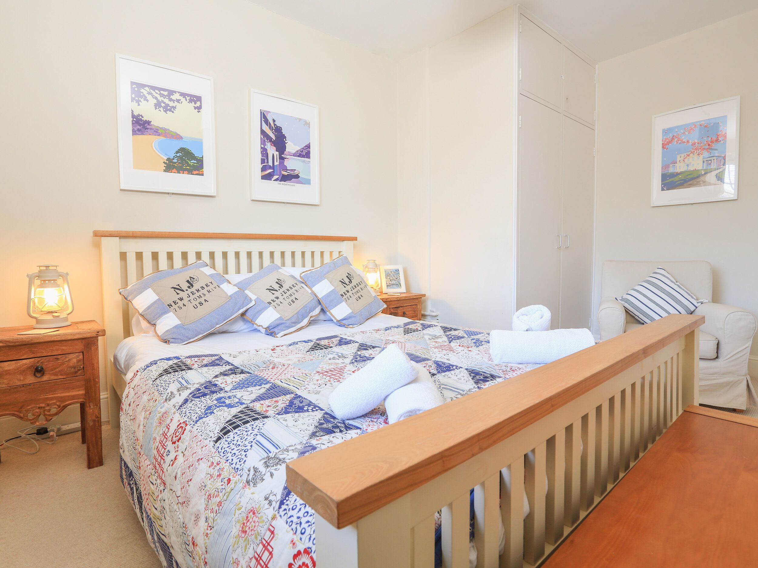 Een net opgemaakt bed met een sprei, kussens en een bedlampje. Er staat een stoel met een gestreept kussen en een foto aan de muur.