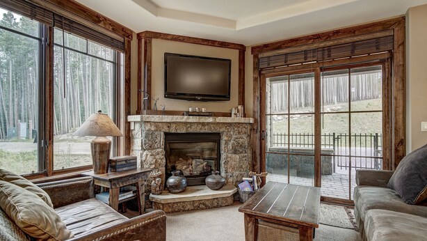 A cozy living room with a stone fireplace, a flat-screen TV mounted above it, and large windows offering a view of the outdoors. - True Ski-in/ski-out Luxury Condo On Peak 7 1 Bedroom Condo by RedAwning (Breckenridge)