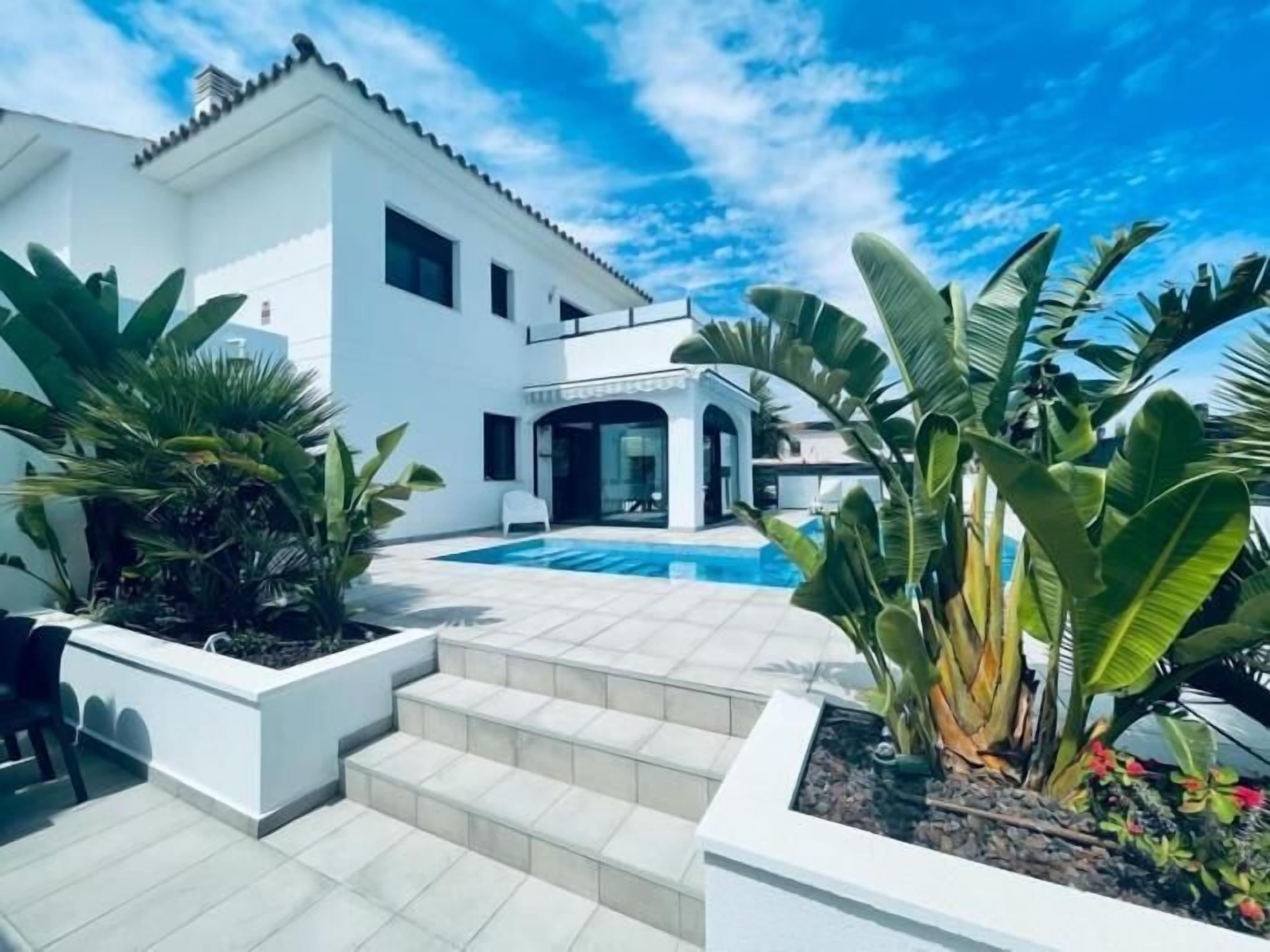 Et moderne hvidt hus med swimmingpool, omgivet af tropiske planter og en klar blå himmel.