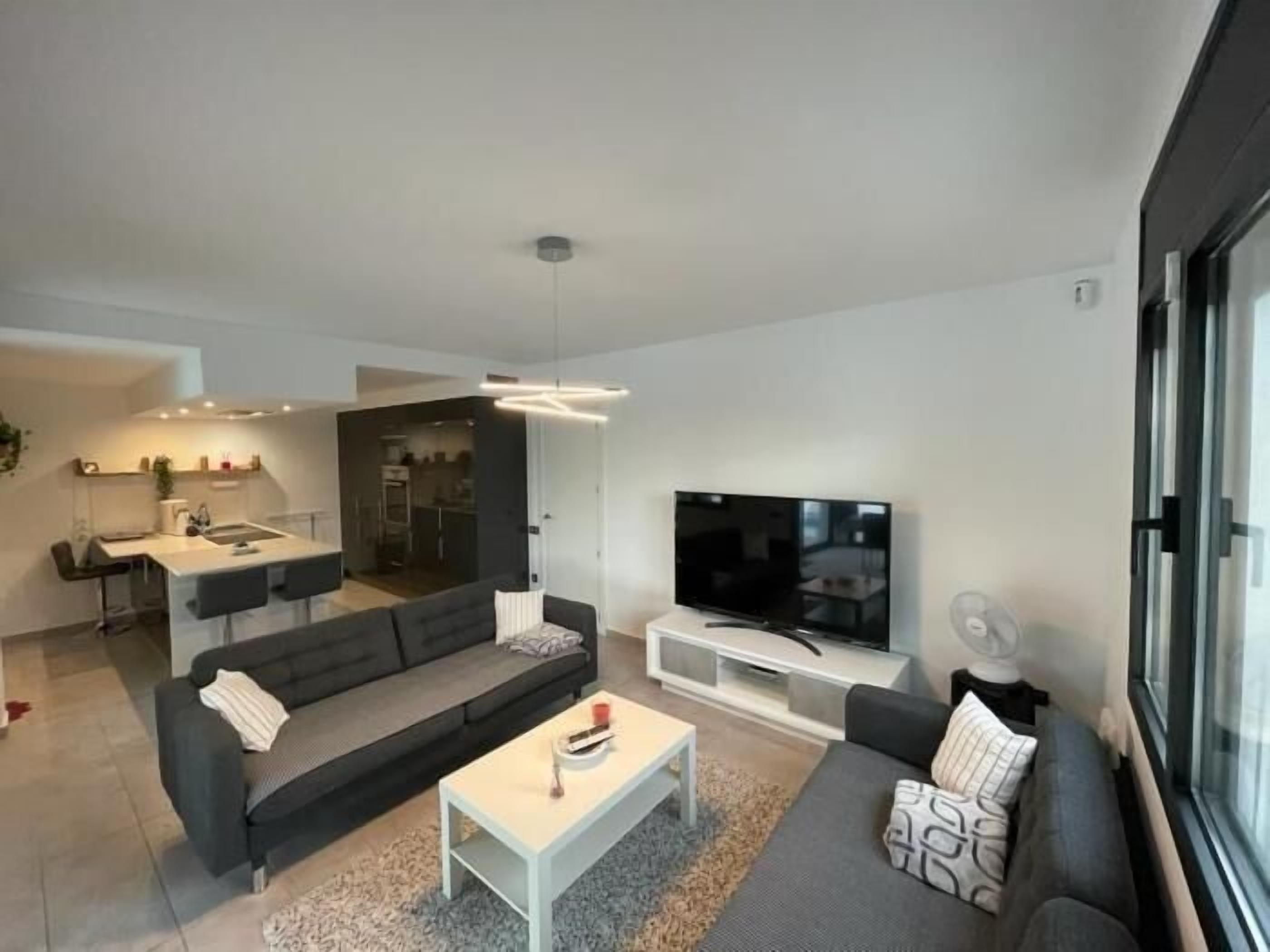 Una sala de estar moderna con un sofá gris, una mesa de centro blanca y un televisor sobre un mueble bajo. Se observa una zona de cocina a través de una puerta abierta.