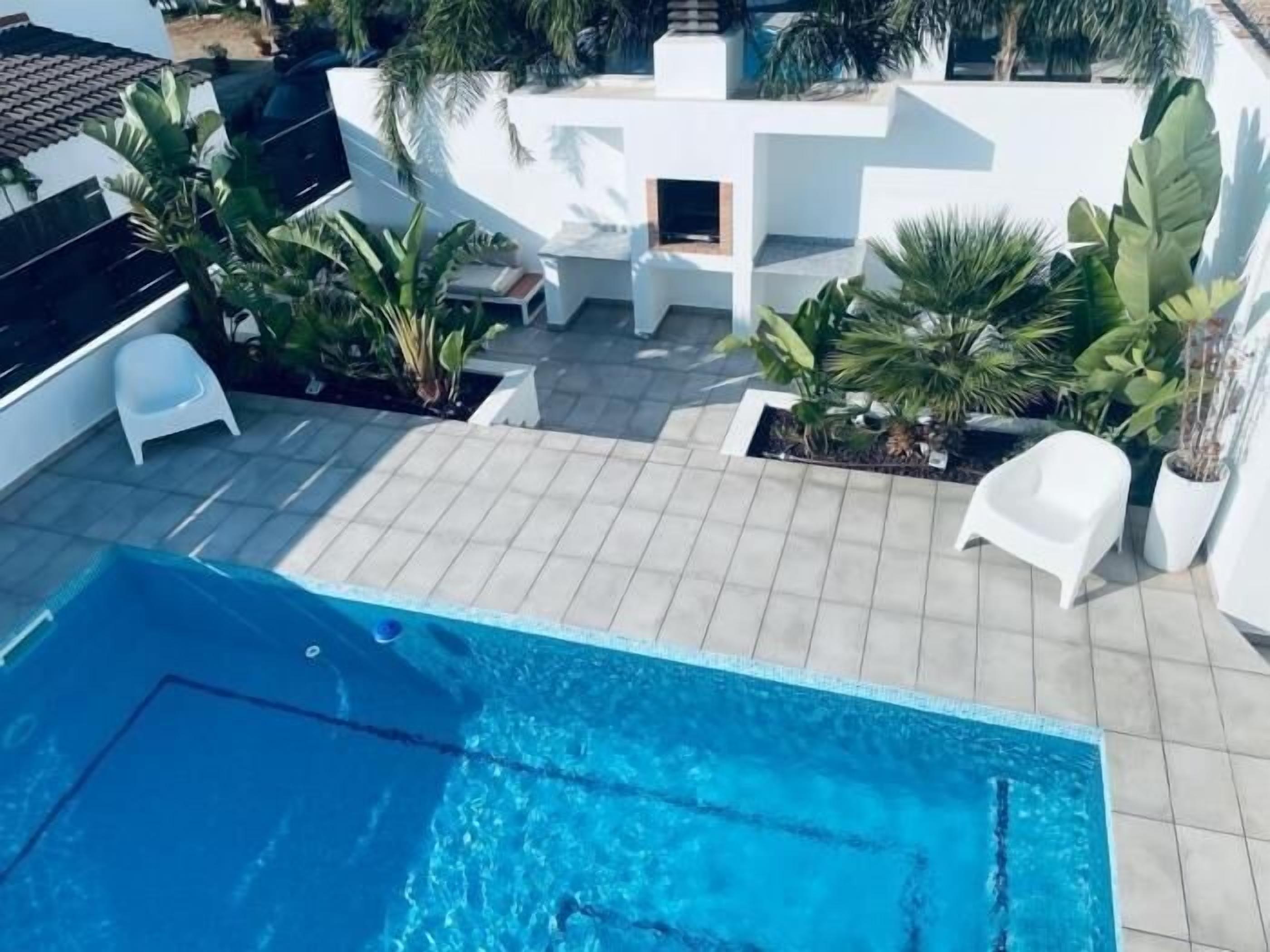 A rooftop swimming pool with tiled flooring, surrounded by potted plants and white chairs.