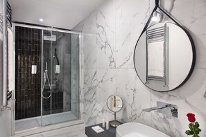 A modern bathroom with a glass-enclosed shower, a round mirror, a white bathtub, and a rose decoration. - Fellini Design Suites (Rome)