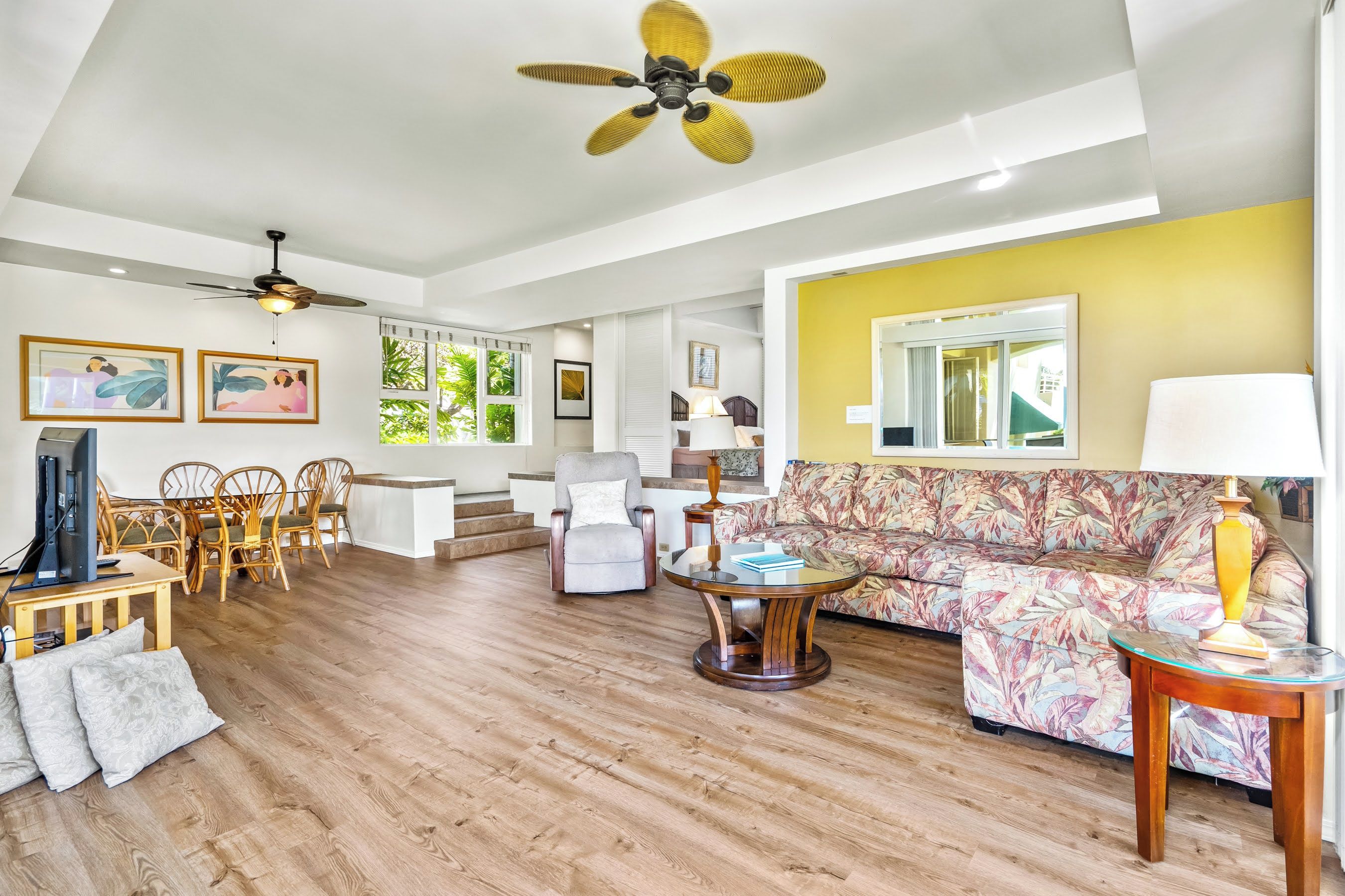 A spacious living room with a wooden floor, a sofa, a coffee table, a TV, and a ceiling fan.