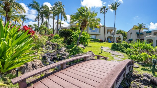 A wooden bridge leading to a resort with palm trees, a small waterfall, and modern buildings in the background. - Palms at Wailea #1207 Ground Floor Unit, Near Complex Pool and Shared Hot Tub (Wailea)