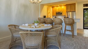 A dining area with wicker chairs and a round table, a modern kitchen with wooden cabinets, and a decorative ceiling light. - Summer Deal Wailea Condo with Ocean Views Golf Course and Pools (Wailea)