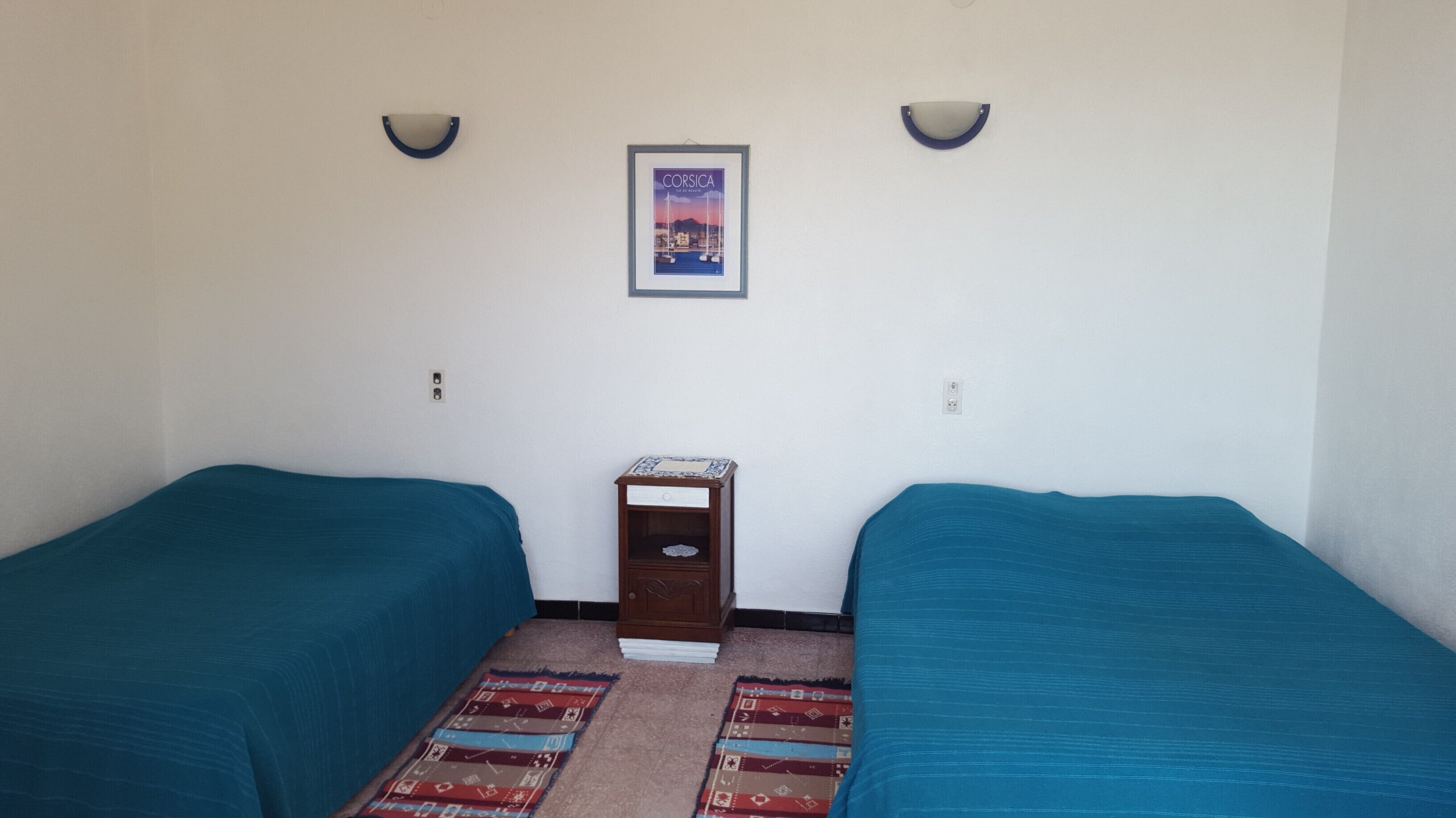 Una habitación con dos camas individuales, una mesita de noche de madera, un cuadro en la pared y dos lámparas de pared.