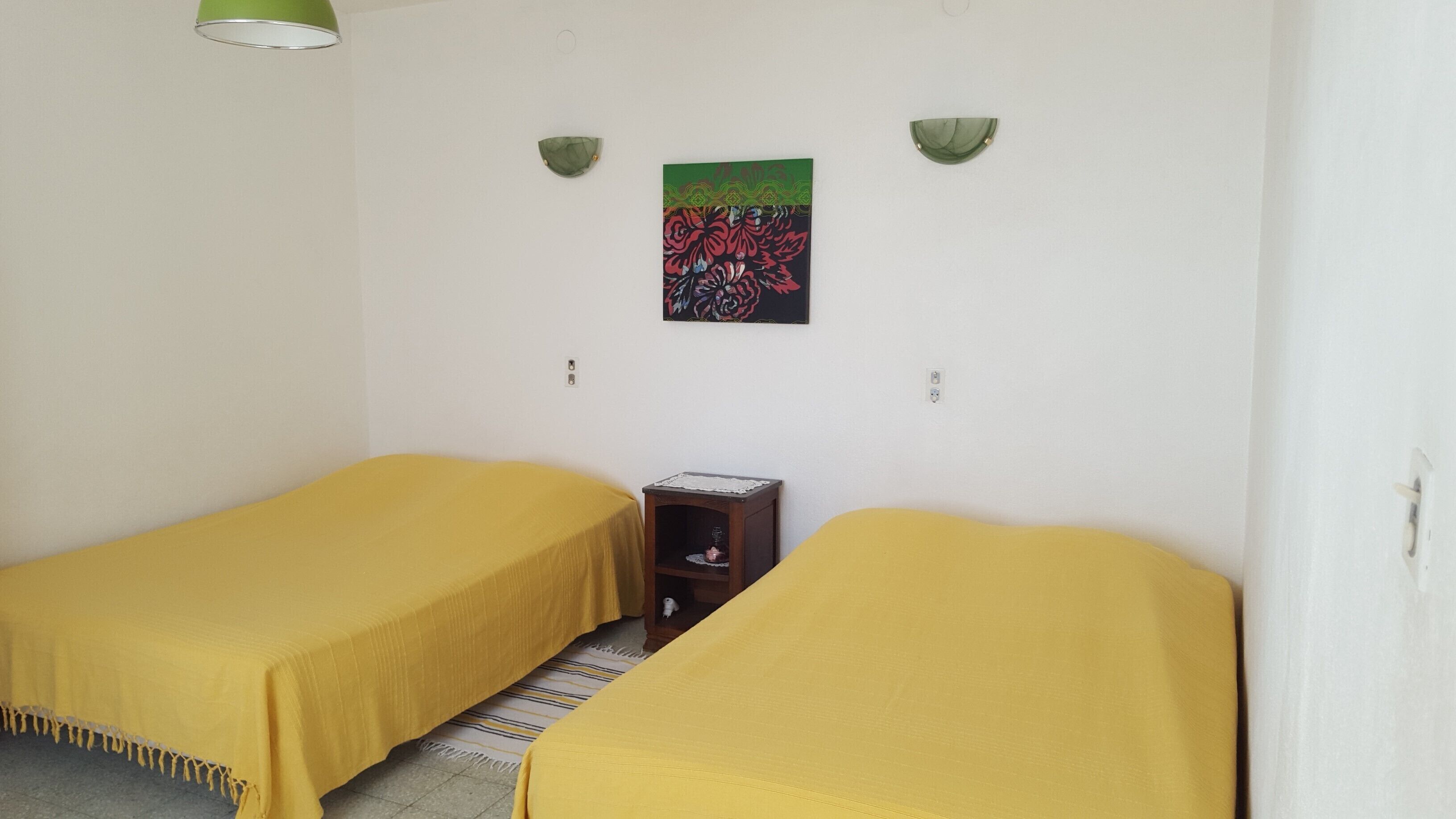A room with two single beds, each covered with a yellow bedspread. There is a small wooden nightstand with a lamp between the beds. A colorful floral artwork is hung on the wall above the beds.