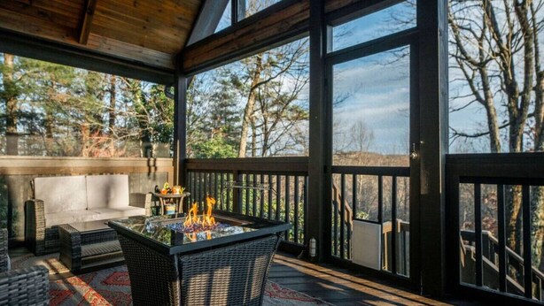 A covered outdoor seating area with a fire feature, wicker furniture, and a view of trees.
