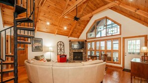 A spacious living room with a large L-shaped sectional sofa, a stone fireplace, a wooden ceiling, and a spiral staircase.
