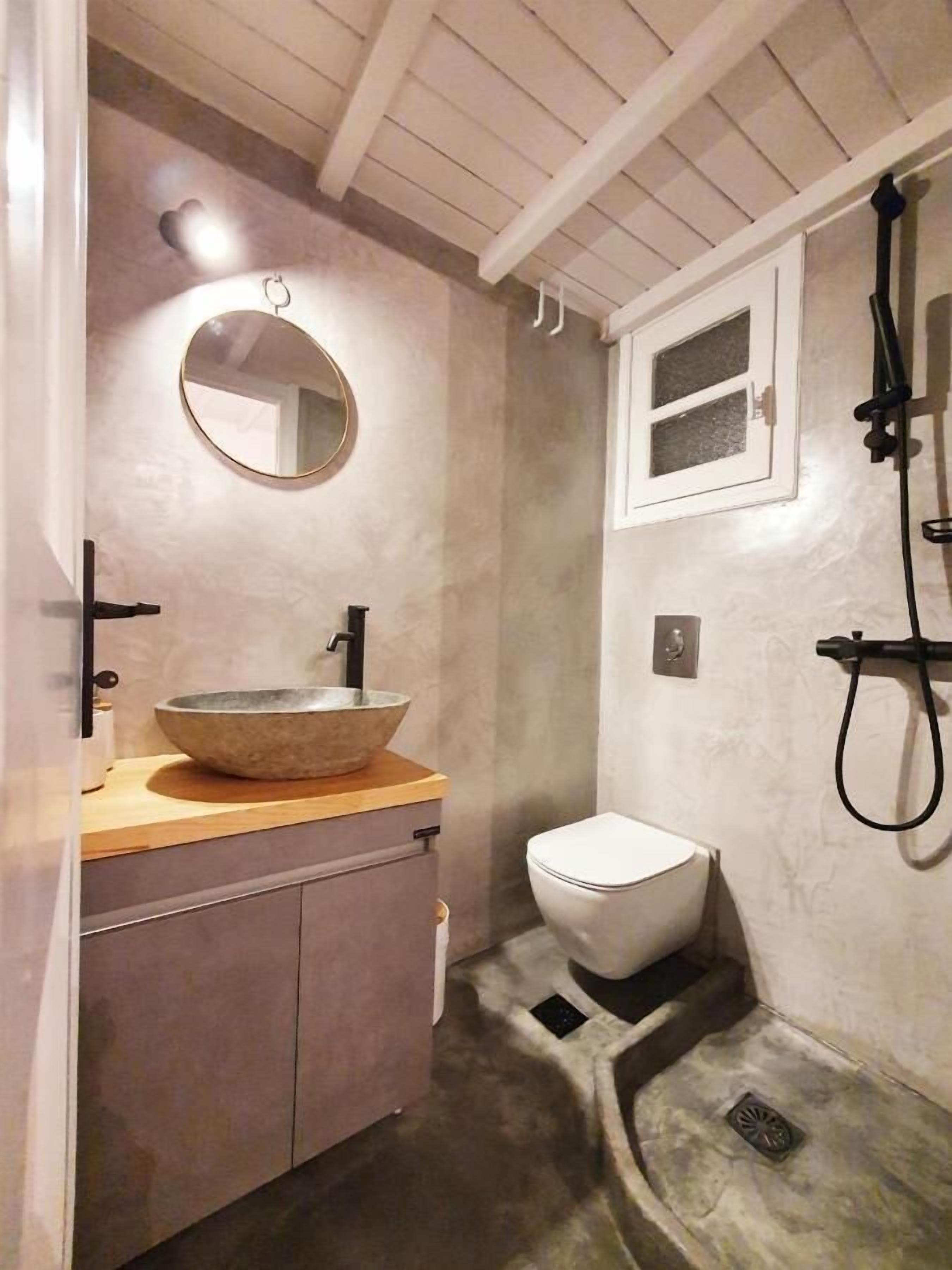 Un baño con un lavamanos de piedra, un espejo redondo, un inodoro y una ducha con manguera.