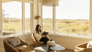 A room with large windows, a reading chair, a small table, and a lamp.