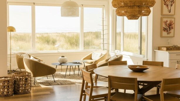 A spacious room with a large dining table, comfortable seating, and large windows offering a view of the beach.