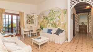 Une pièce avec un canapé blanc, une table basse, une table à manger avec des chaises et une fresque représentant des plantes tropicales.