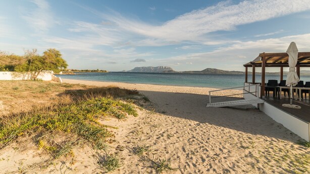 Une plage avec une terrasse en bois, des sièges en plein air et une vue sur la mer et les montagnes lointaines.