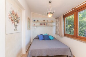Une chambre à coucher avec un lit, une fenêtre, un lustre, des étagères remplies de livres et d’objets décoratifs, et un tableau accroché au mur.