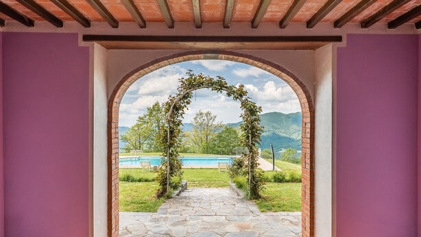 Ein rundbogiger Durchgang, der zu einem Steinweg mit einem Pool und Bergen im Hintergrund führt.