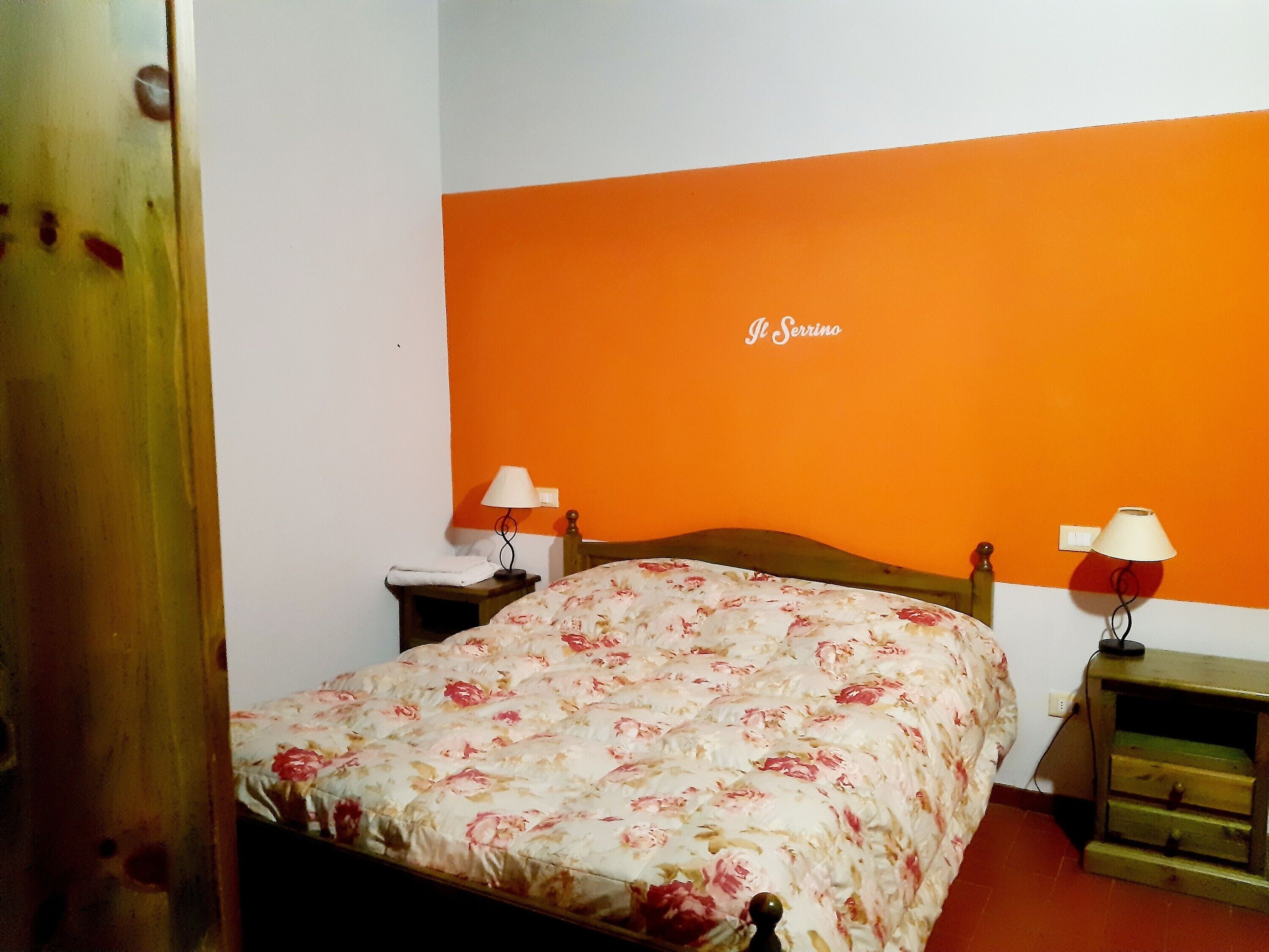 Une chambre à coucher avec une couvre-lit à motifs floraux, deux tables de chevet avec des lampes, un mur d’accent orange et une porte en bois.