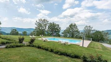 Ein Pool mit Liegestühlen, umgeben von einer Rasenfläche und Bäumen, mit Bergen im Hintergrund.