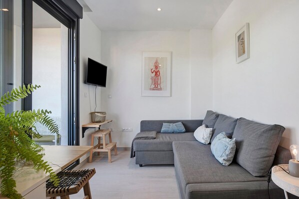 A modern living room with a grey sofa, a small wooden table, a TV mounted on the wall, and a framed artwork.