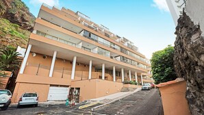 Un edificio de varios pisos con balcones, zona de estacionamiento y un paisaje montañoso de fondo.