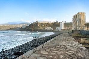 A coastal walkway with a stone path, ocean waves, and buildings in the background.
