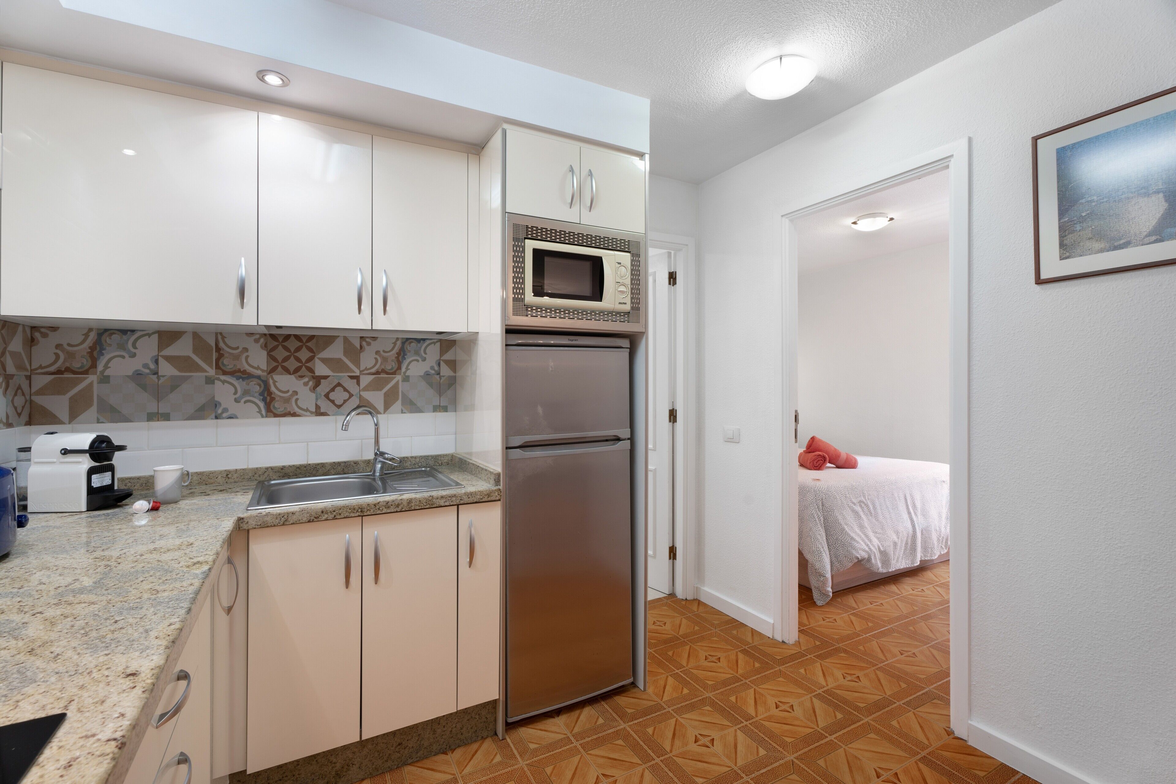 Cocina compacta con refrigerador, microondas y pileta, que da a un dormitorio con cama y un cuadro.