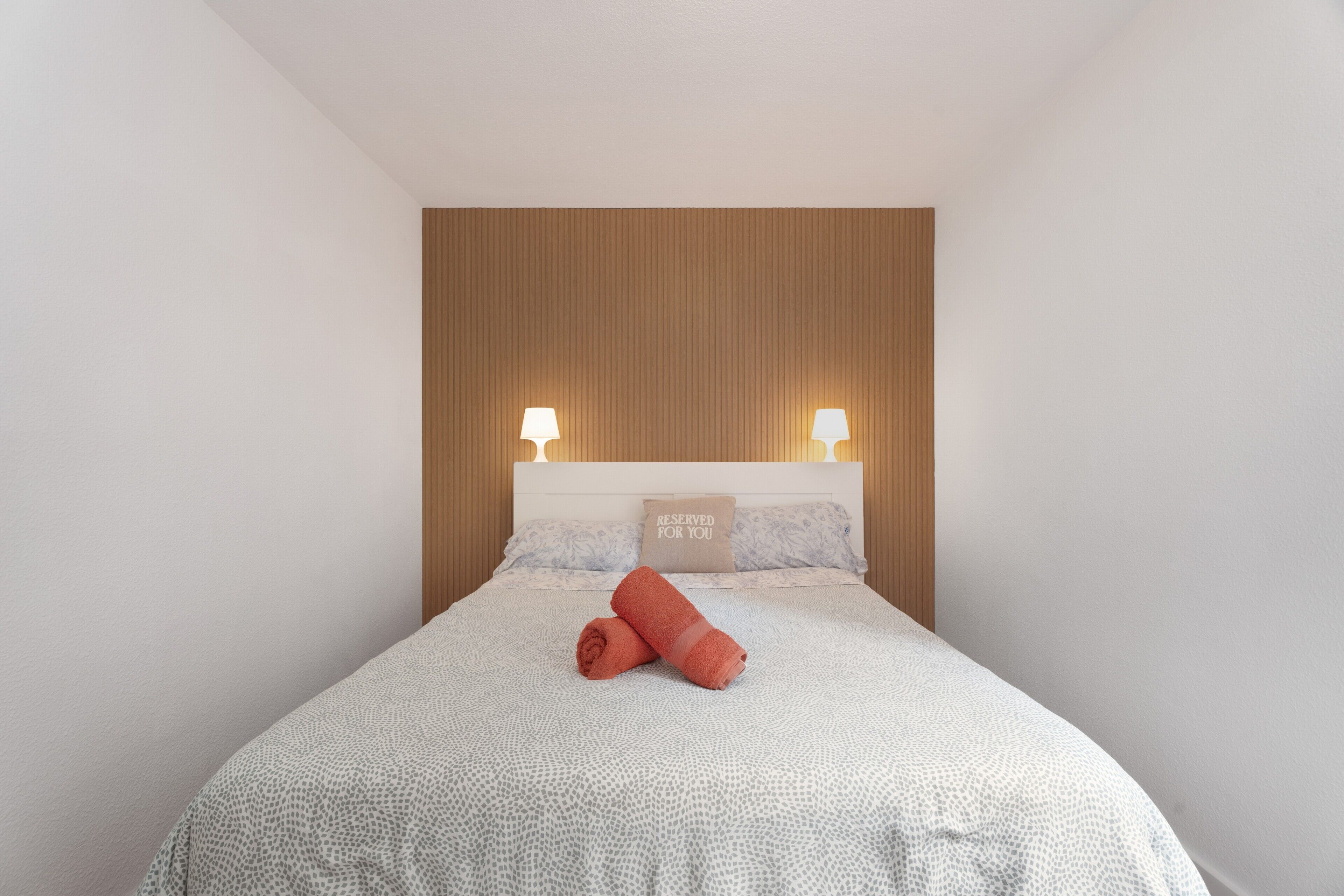 A neatly made bed with a patterned bedspread, two bedside lamps, and a pillow with 'RESERVED FOR YOU' text.