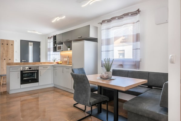 A modern kitchen with built-in appliances, a dining area with a wooden table and grey upholstered seating, and a window with sheer curtains.
