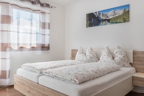 A double bed with floral bedding, a wooden headboard, a nightstand, a wall-mounted painting of a mountain landscape, and a window with a striped curtain.