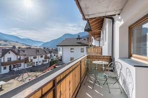 A balcony with a view of a village, a table with chairs, and a window overlooking the mountains.