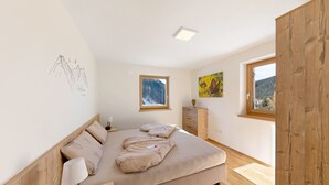 Ein Schlafzimmer mit Bett, Nachttisch, Lampe, Kommode und einem Fenster mit Bergblick.