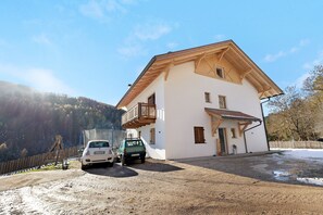 Una casa de dos pisos con balcón, un camino de entrada con dos coches y una montaña boscosa al fondo.