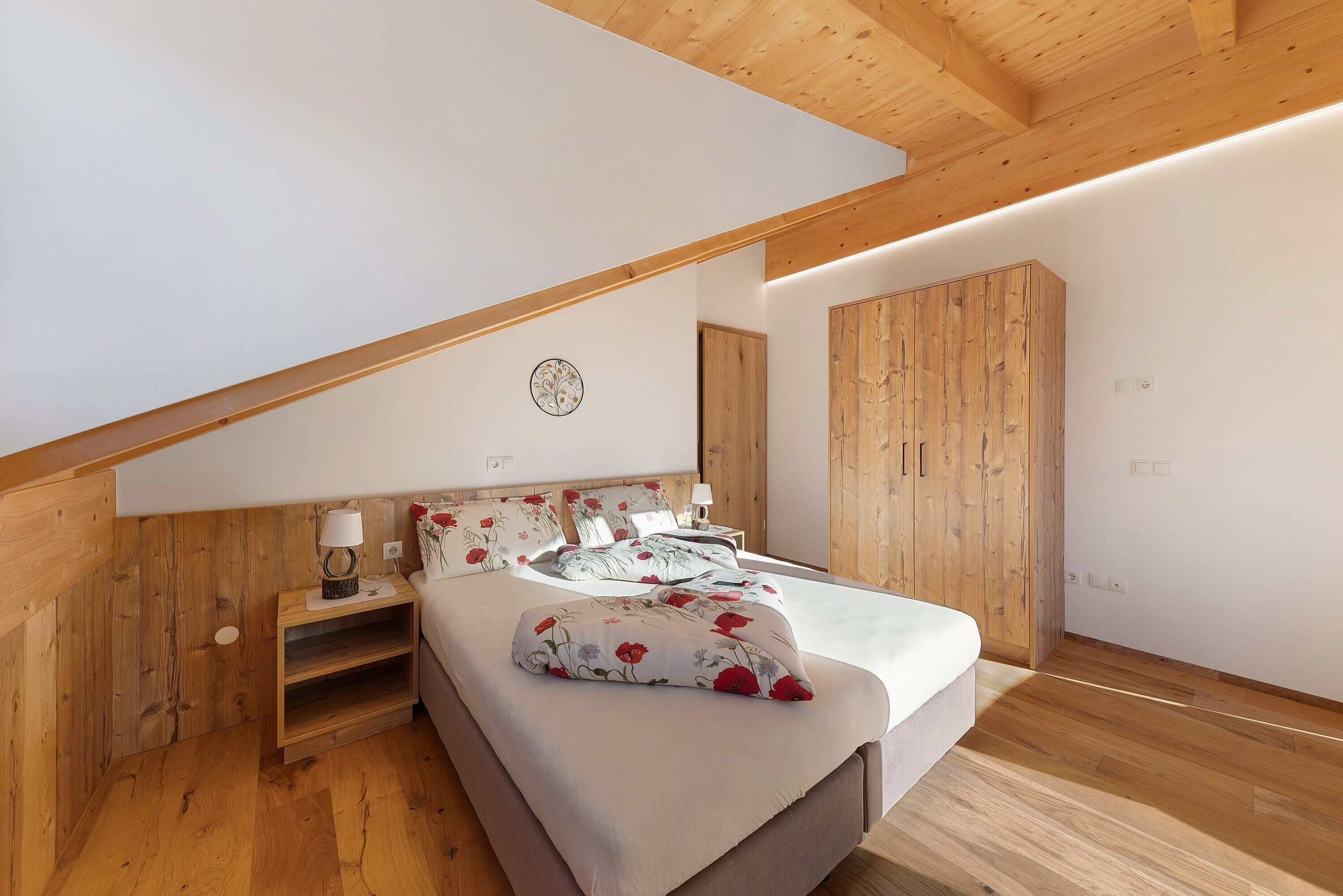 Een slaapkamer met schuine wanden, houten lambrisering, een bed met bloemenbedrukking, een nachtkastje en een bedtafel.