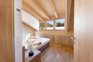 A bedroom with a wooden ceiling, a bed with floral bedding, a bedside table with a lamp, and a window with a view of trees.