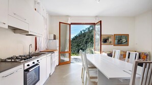 A modern kitchen with a gas stove, oven, and white cabinetry. There is a dining area with a white table and chairs, and a view of a forest through an open door.