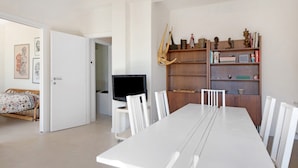 A modern living room with a dining table, white chairs, a television, and a bookshelf with decorative items.