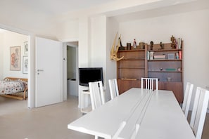 Una sala de estar moderna con una mesa de comedor, sillas blancas, un televisor y una estantería con objetos decorativos.
