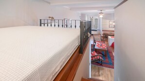 Une chambre à coucher avec un lit, un escalier en bois, une petite table recouverte d’une nappe rouge et une vue sur un couloir.