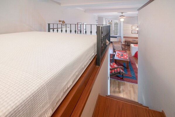 A bedroom with a bed, a wooden staircase, a small table with a red tablecloth, and a view of a hallway.