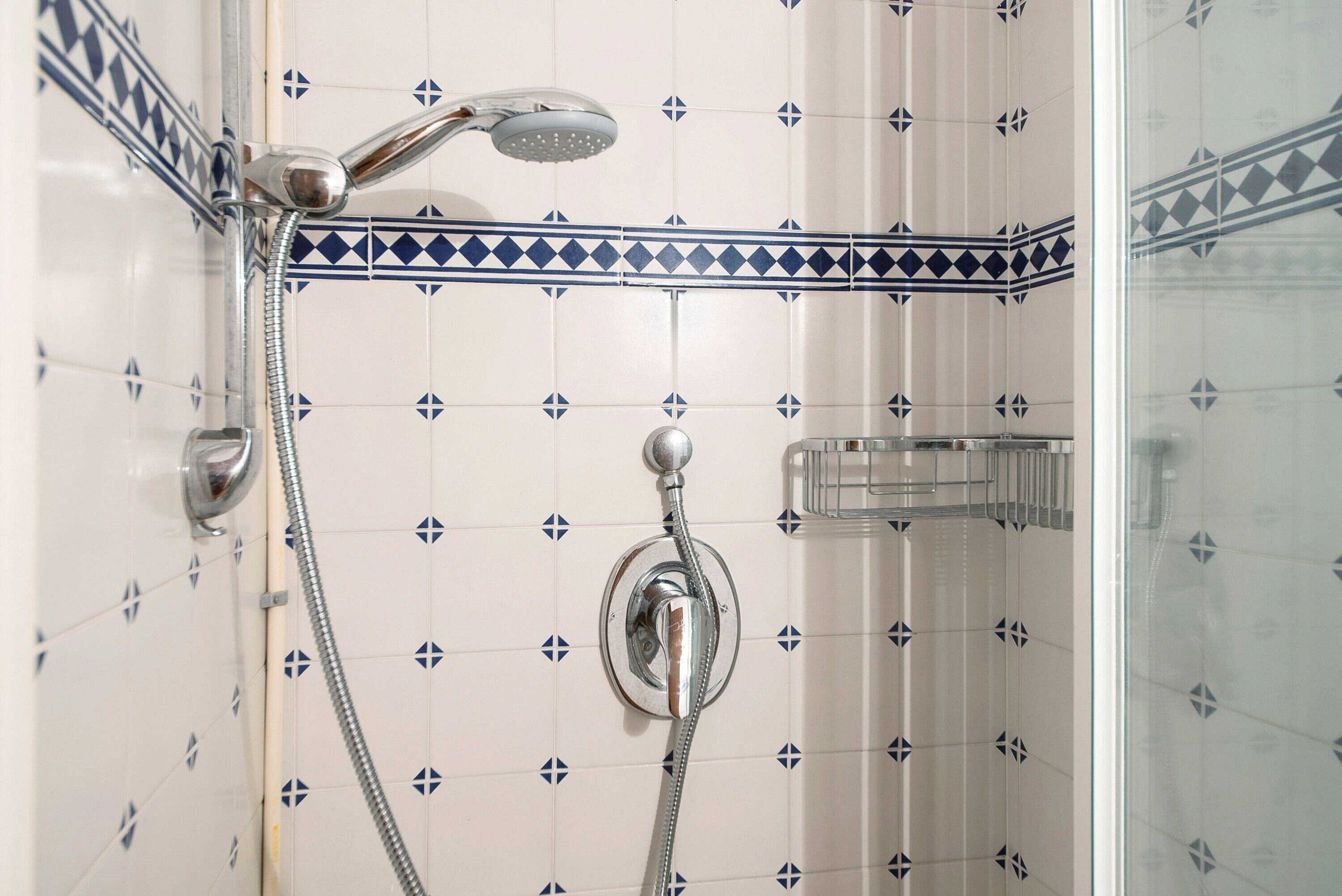 Een douche met een handdouche en een vastgeschroefde douchekop, een glazen douchemandje, en een betegelde muur met een ruitmotief.