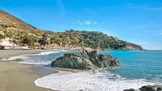 Une plage côtière bordée de formations rocheuses, avec une eau d’un bleu limpide et une colline parsemée de bâtiments.